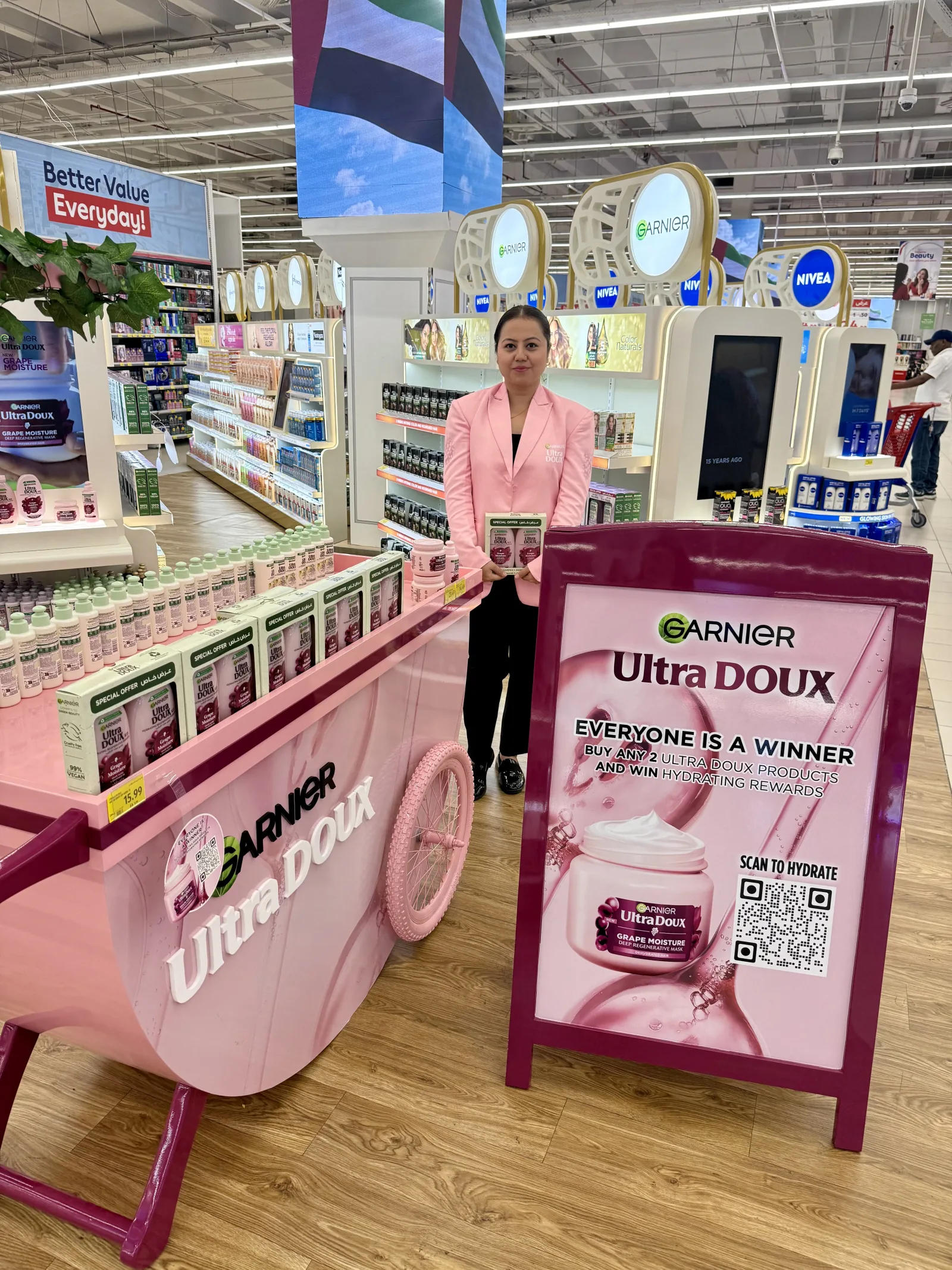 A beauty advisor beside a pink Garnier Ultra Doux sampling cart in a UAE hypermarket aisle.