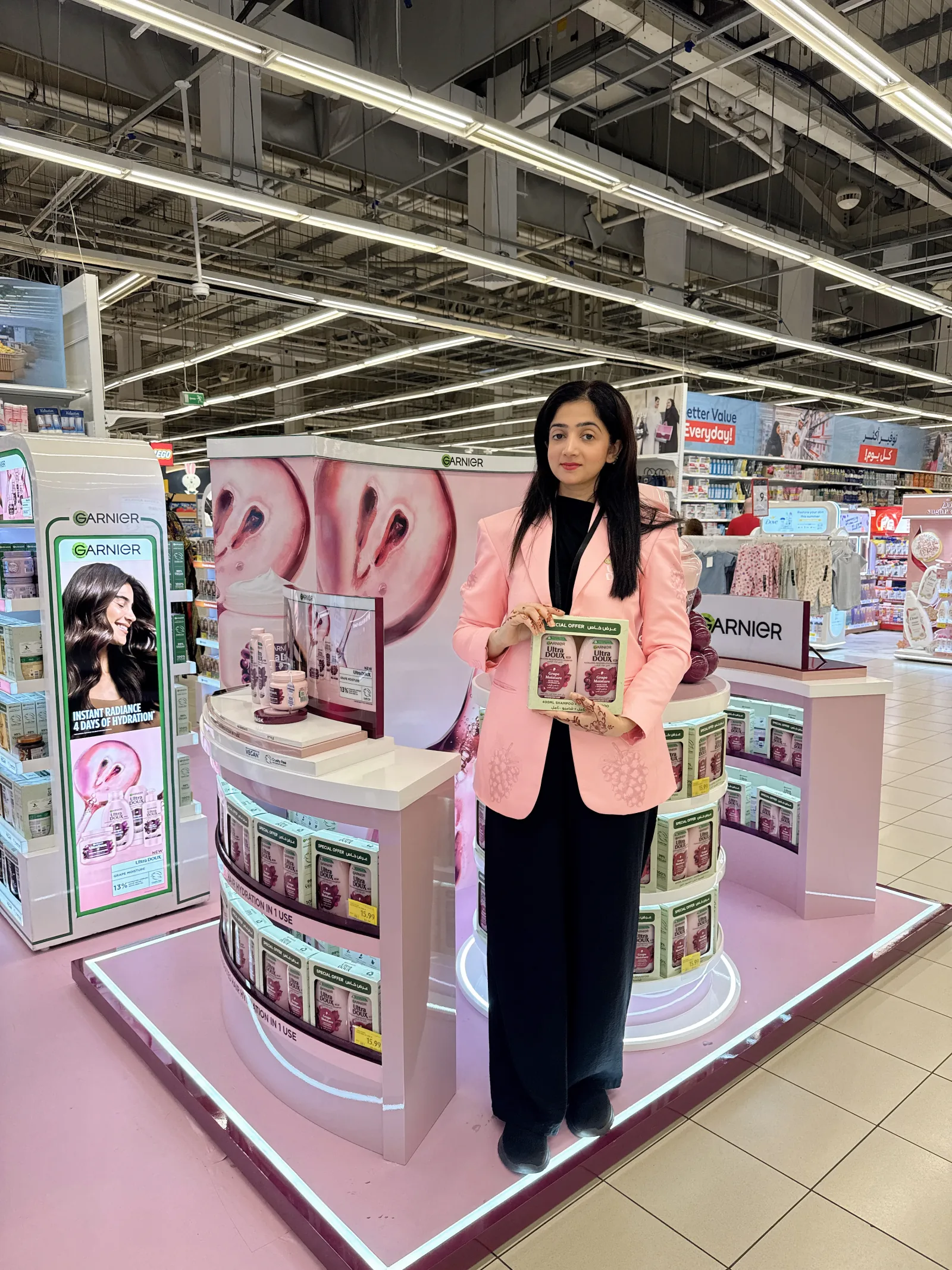 A beauty advisor holding a leaflet in front of a large pink Garnier Ultra Doux in-store display island.