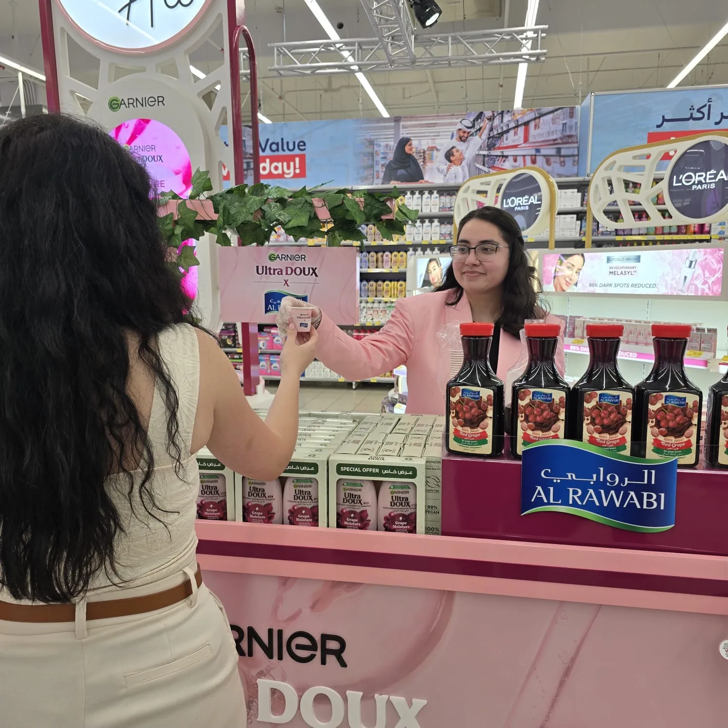 A beauty advisor handing a Garnier Ultra Doux product to a shopper across the Al Rawabi-branded activation counter.