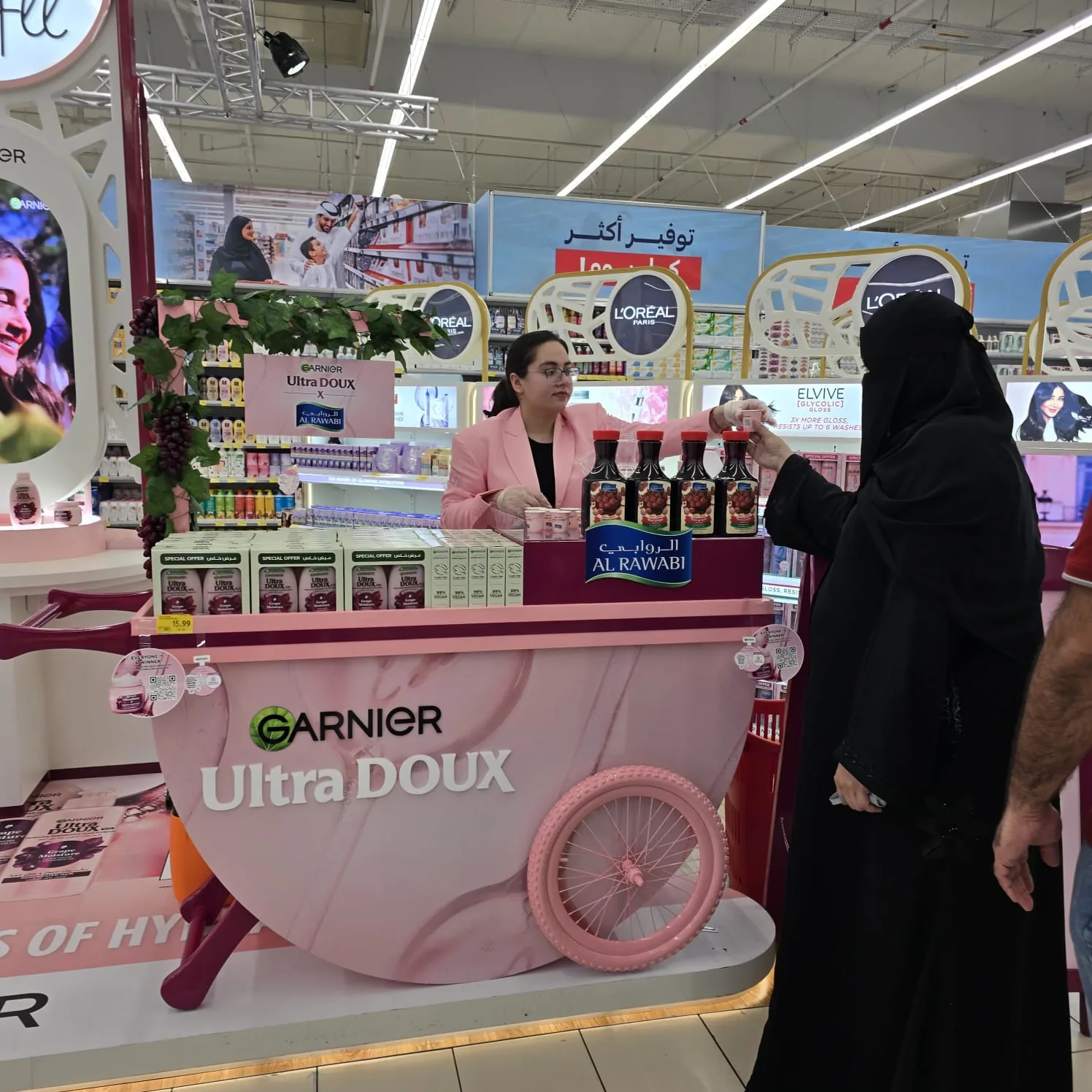 Shoppers being served at the pink Garnier Ultra Doux cart, Al Rawabi bottles lined up along the counter.