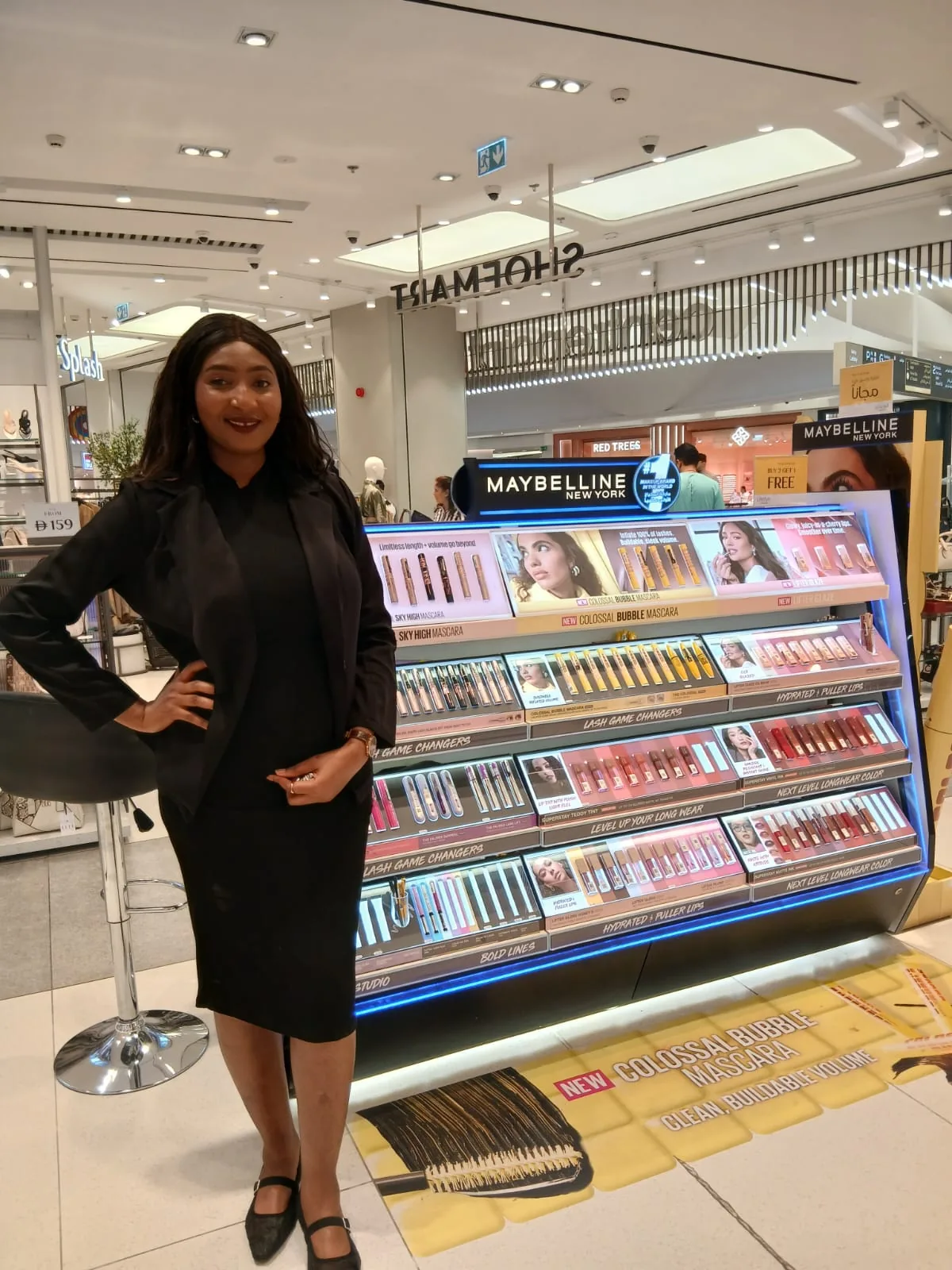 A beauty advisor beside the Maybelline New York gondola in a department-store beauty hall.