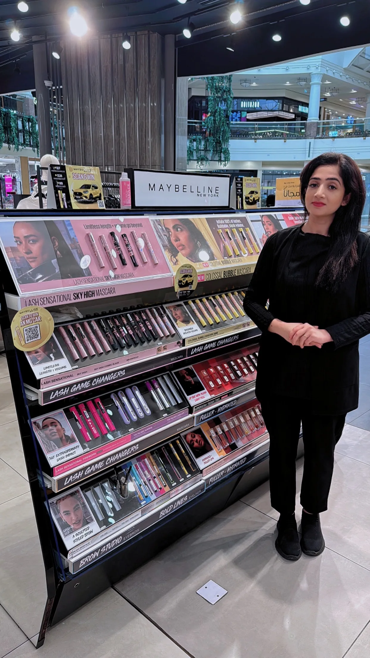 A beauty advisor beside a Maybelline New York display unit stocked with lip and face products.