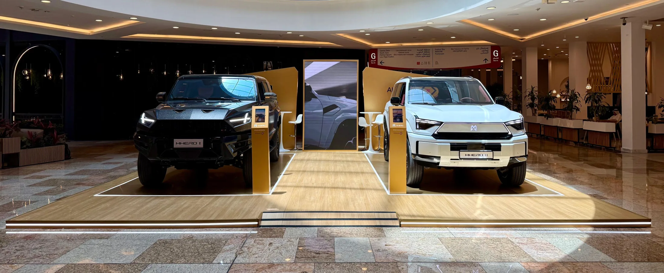The MHERO stand inside Dubai Festival City Mall — the black MHERO I and white MHERO II facing each other across a wood platform with a central screen.