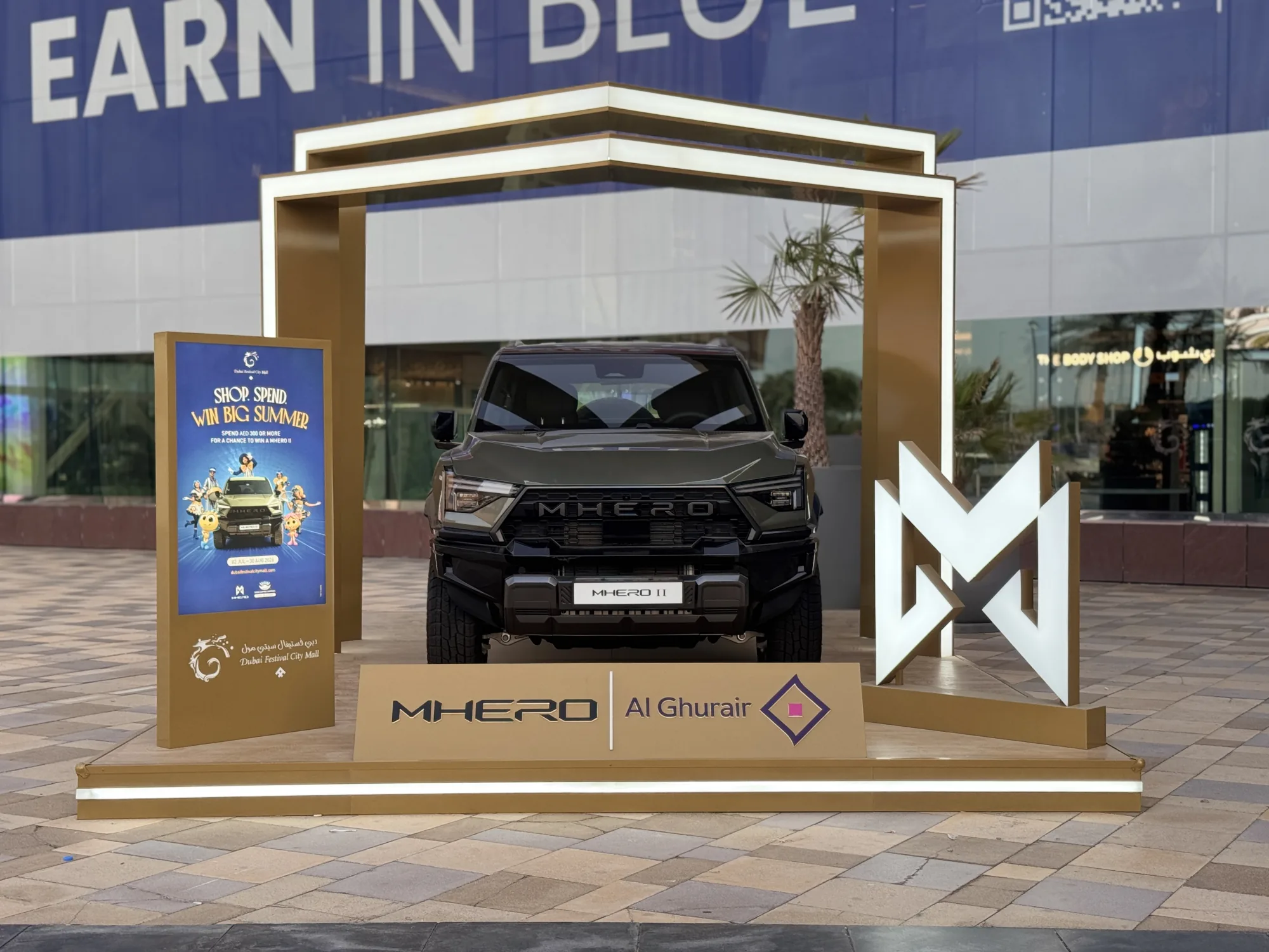 The outdoor MHERO stand — a MHERO II framed by a gold gateway and the illuminated MHERO monogram outside Dubai Festival City Mall.