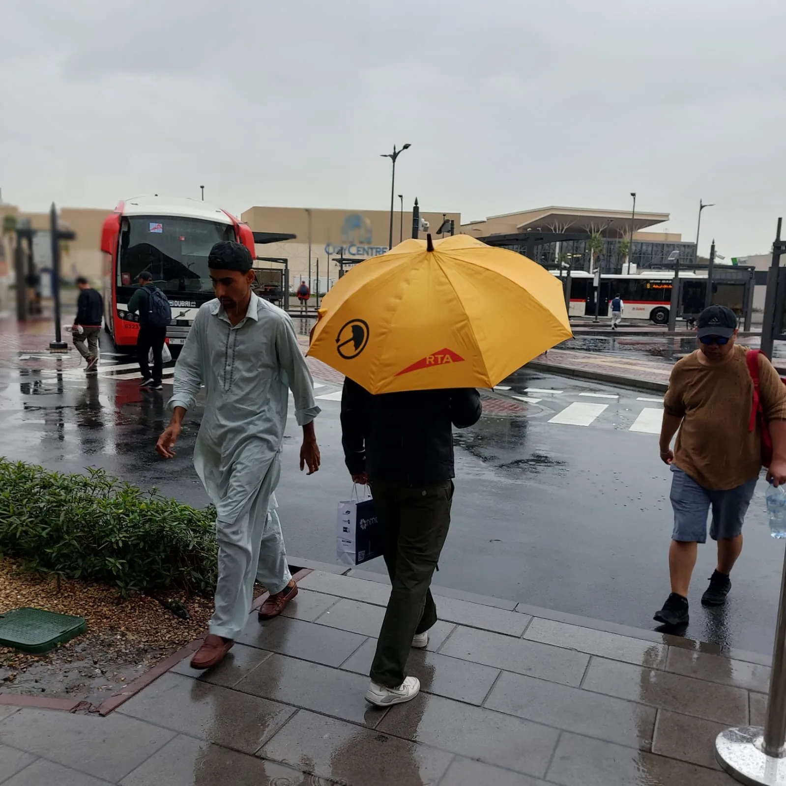 A commuter crossing a rain-soaked Dubai street under a yellow UmbraCity umbrella beside an RTA bus stop.