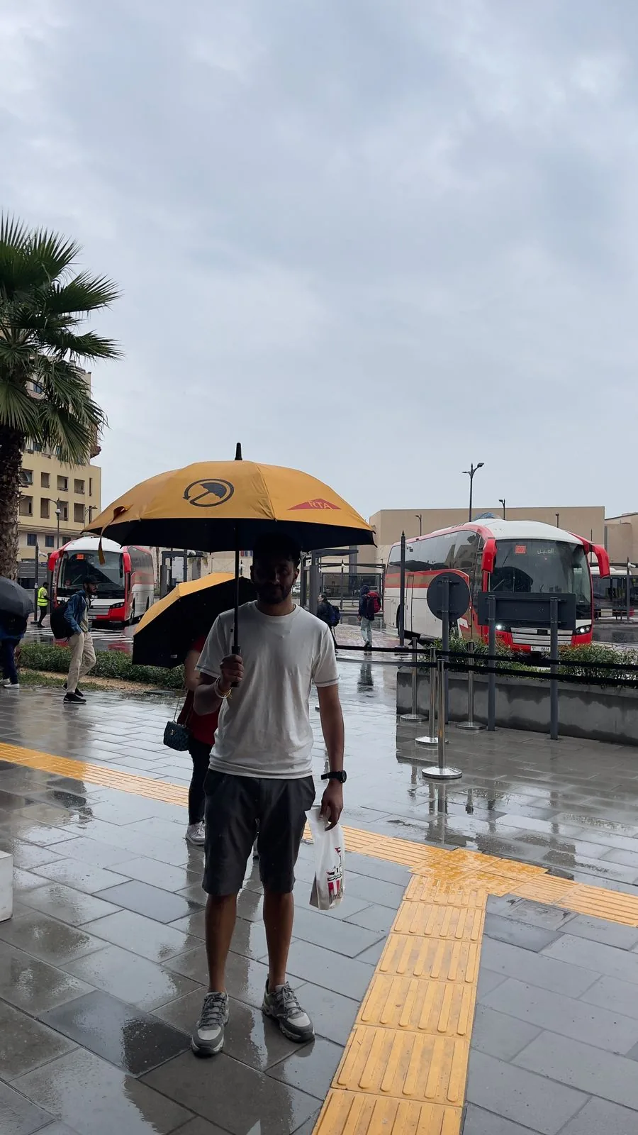 A commuter walking through a Dubai bus station in the rain under a yellow UmbraCity umbrella.