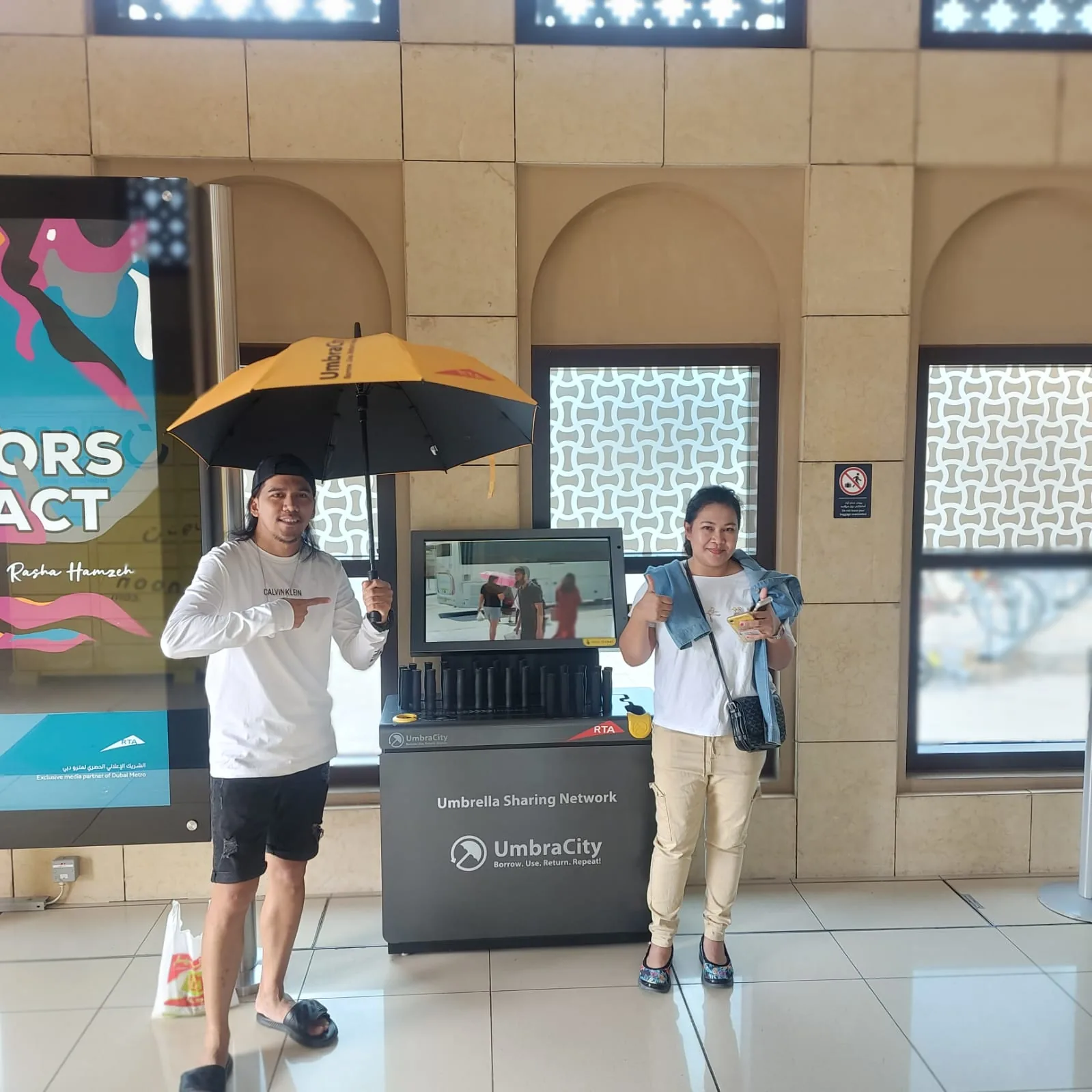 Two commuters giving a thumbs-up beside the UmbraCity umbrella-sharing kiosk at a Dubai Metro station.